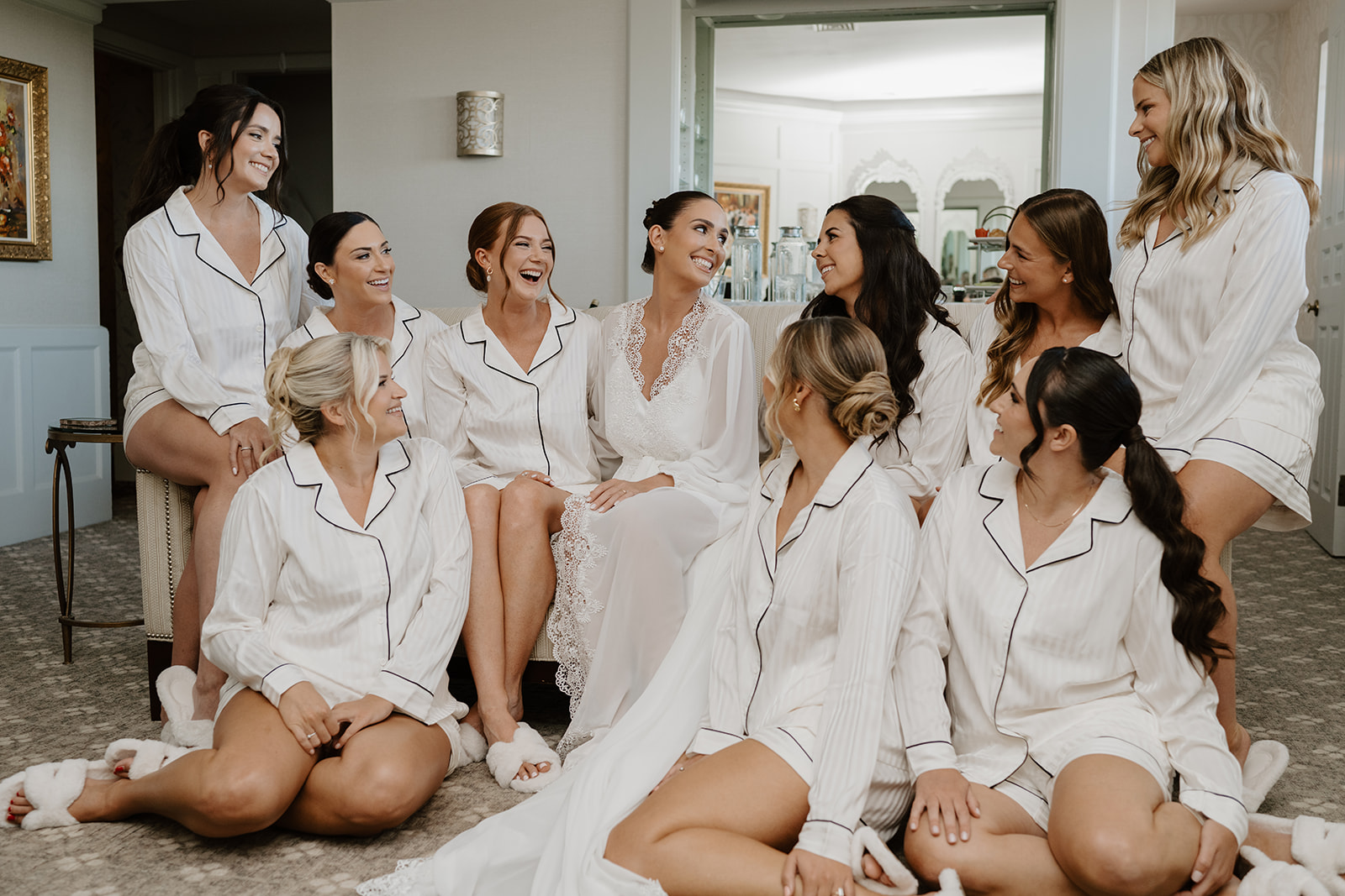 Bride and bridesmaids in matching white pajamas laughing and sharing a happy moment in the bridal suite.